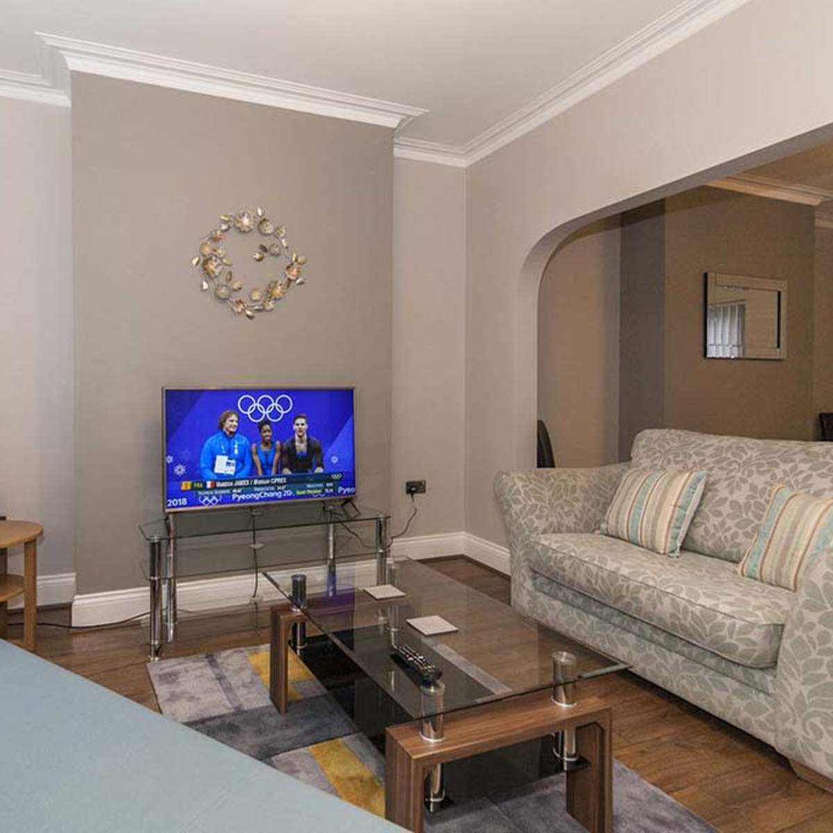 A living room with a light-colored sofa, a patterned couch, a glass coffee table, and a television displaying an Olympic event.