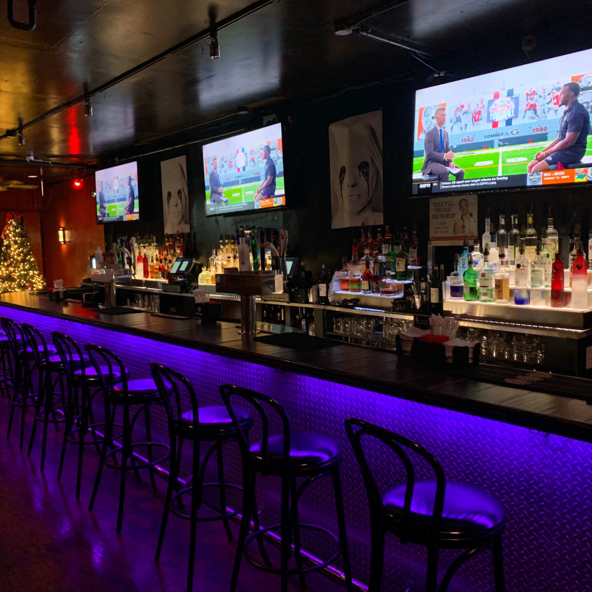 A dimly lit bar with several television screens, a long countertop with bar stools, and a festive Christmas tree in the corner.