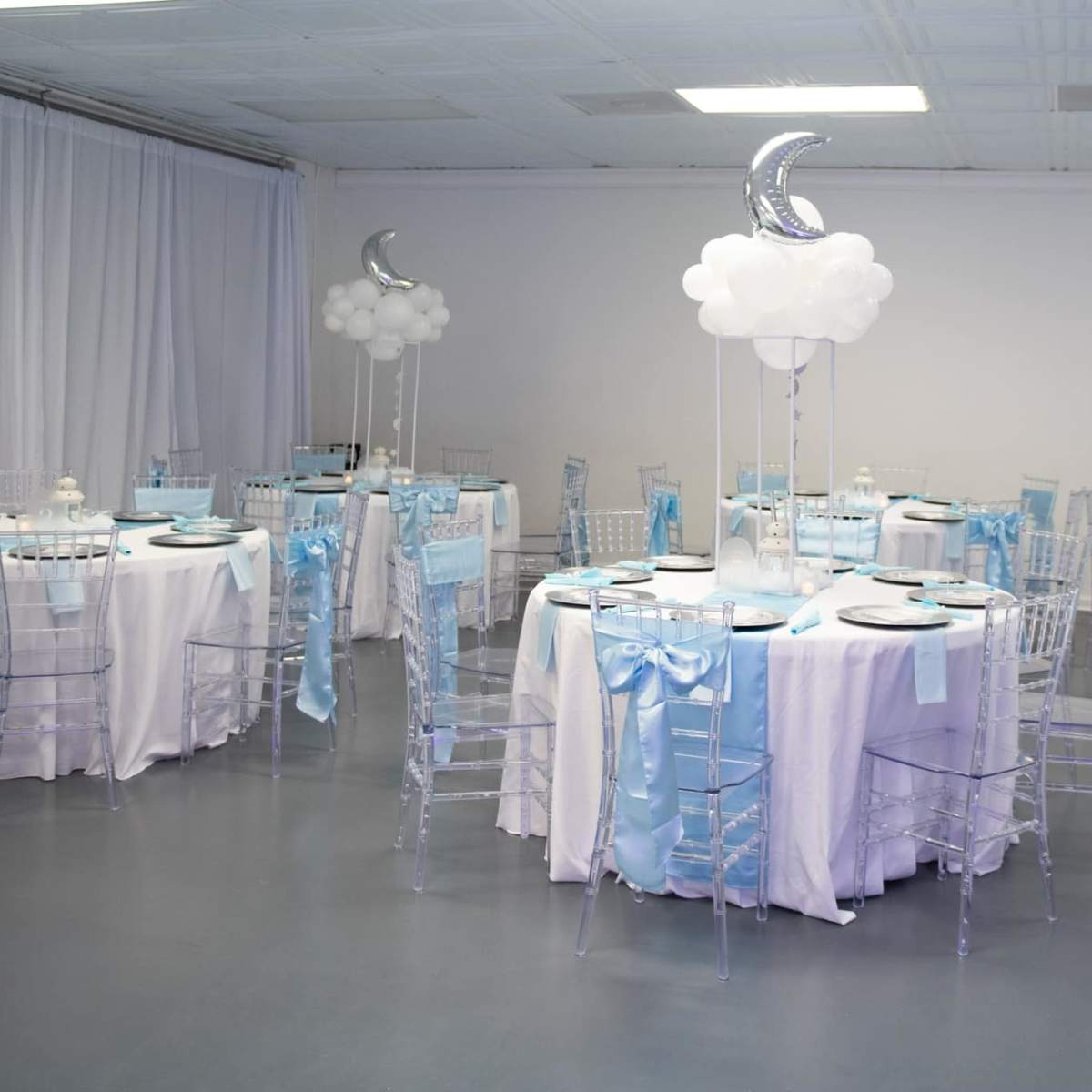 A banquet hall set up for an event, featuring round tables covered with white linens and light blue decorative accents, along with clear chiavari chairs and balloon centerpieces resembling clouds and moons.