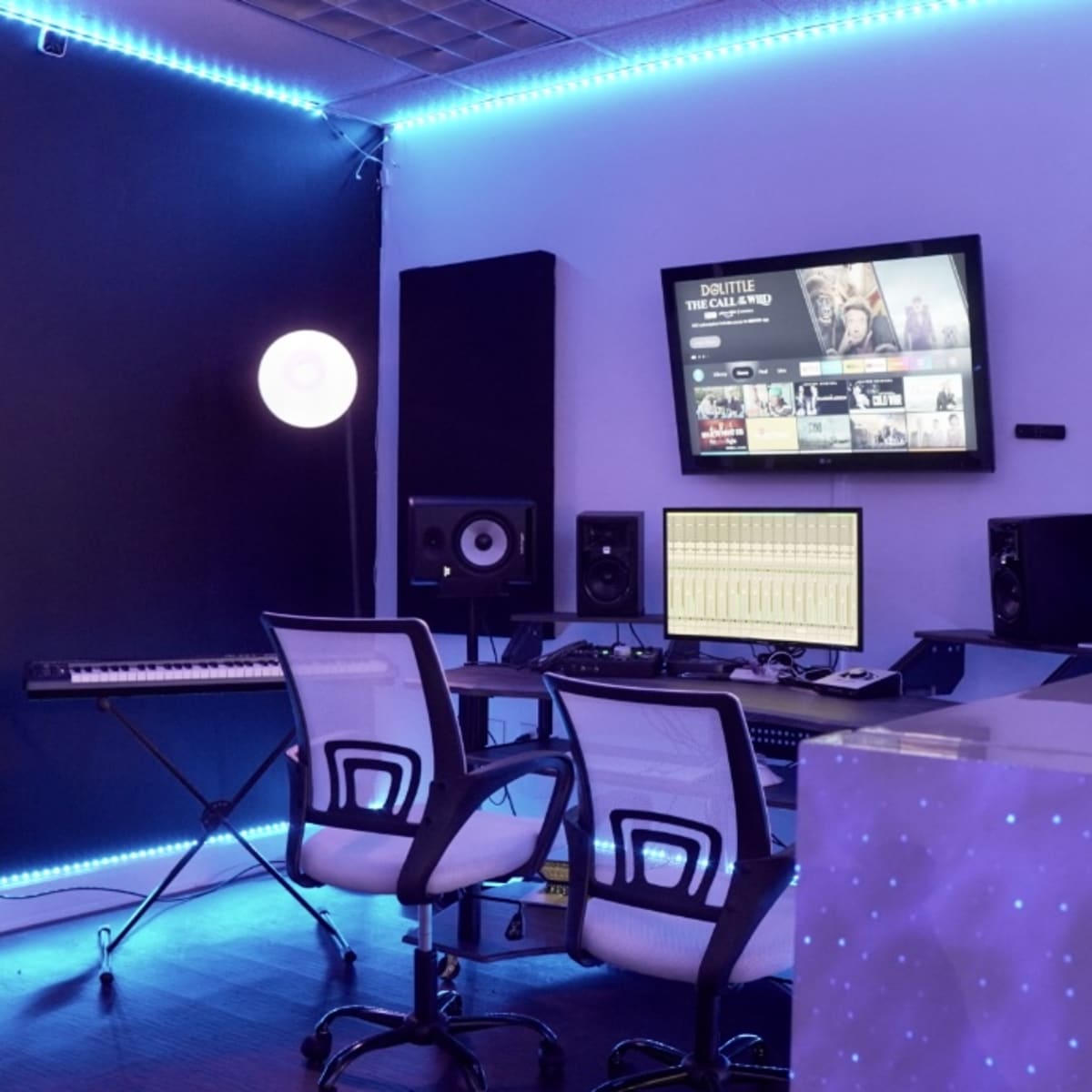 A modern music production studio featuring a desk with two chairs, studio monitors, a keyboard, a large screen mounted on the wall, and ambient LED lighting.