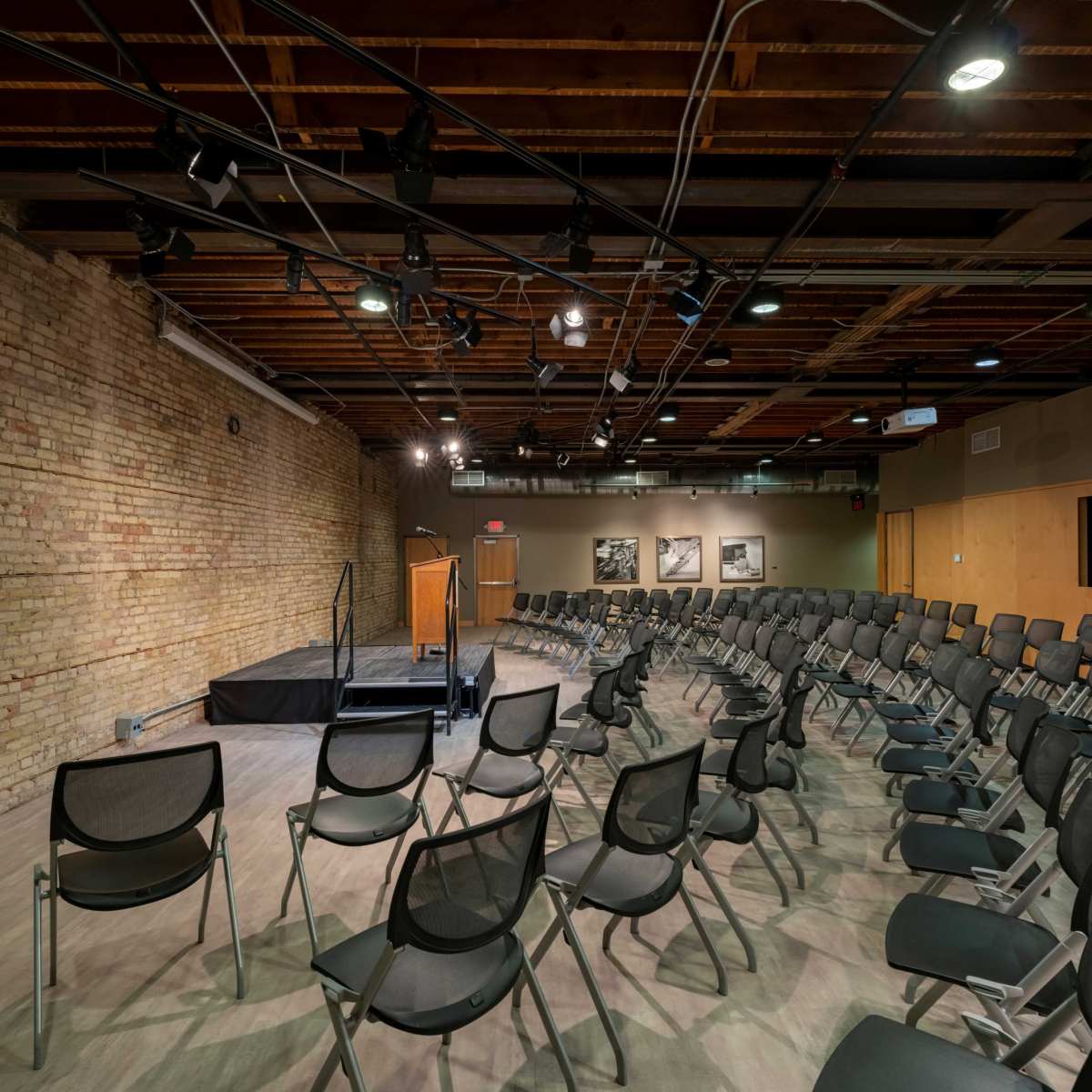 An empty auditorium with rows of chairs facing a small stage, and exposed brick walls.