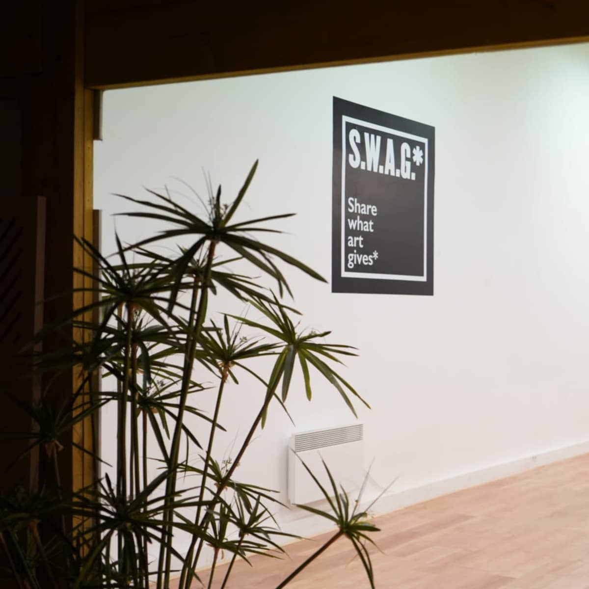 A minimalist interior space featuring a large wall sign that reads "S.W.A.G.: Share What Art Gives," with potted plants in the foreground.