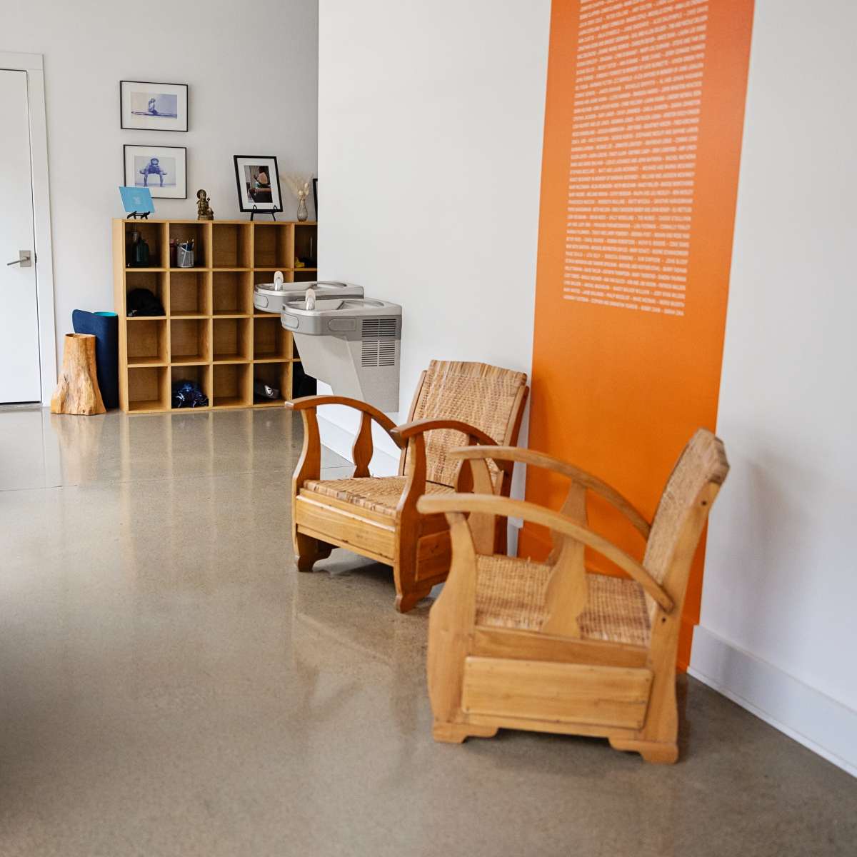 A modern interior space with two wooden chairs, a bookshelf filled with books, a water fountain, and a door leading to another room.