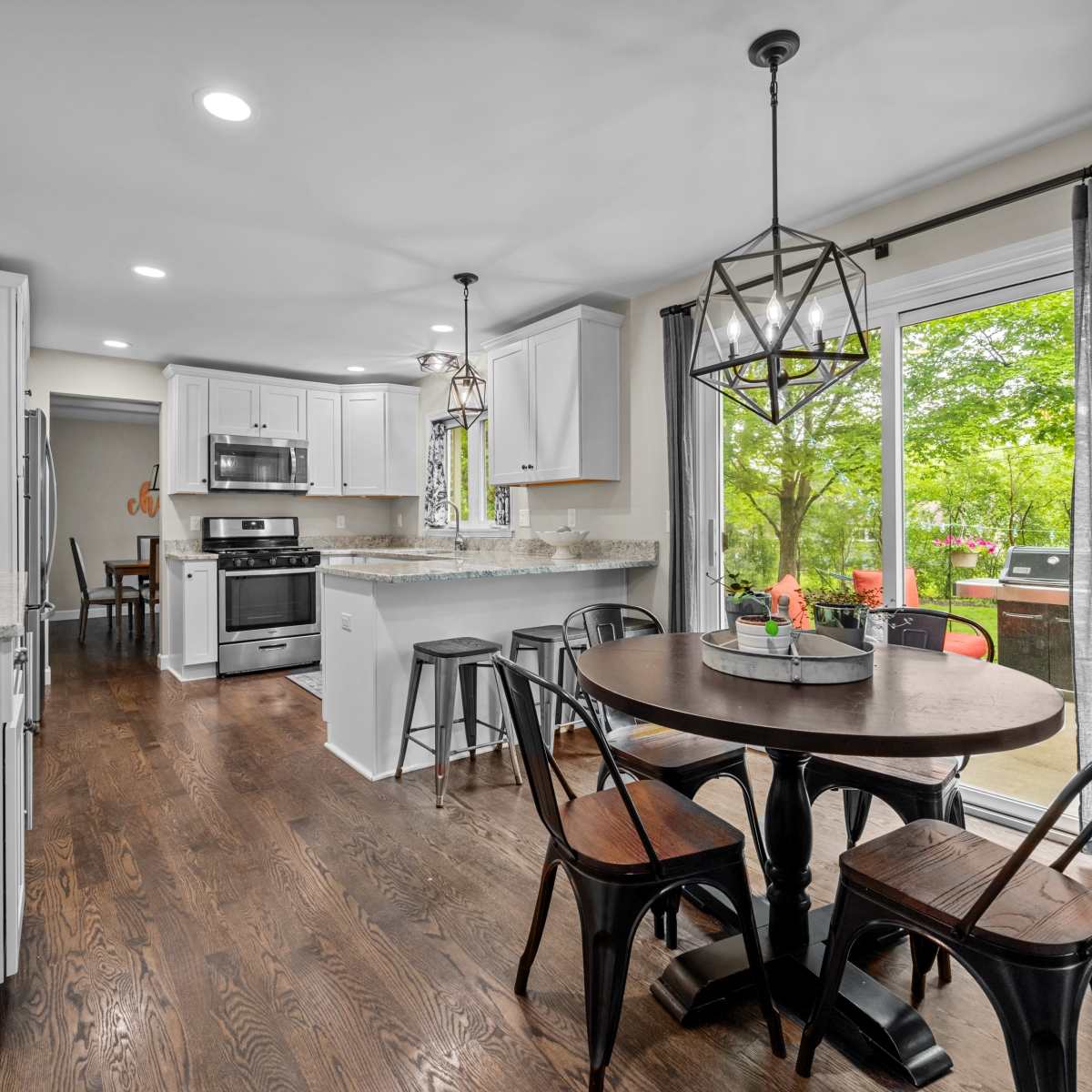 A modern kitchen with white cabinetry, stainless steel appliances, a round dining table, and large windows overlooking a green outdoor area.