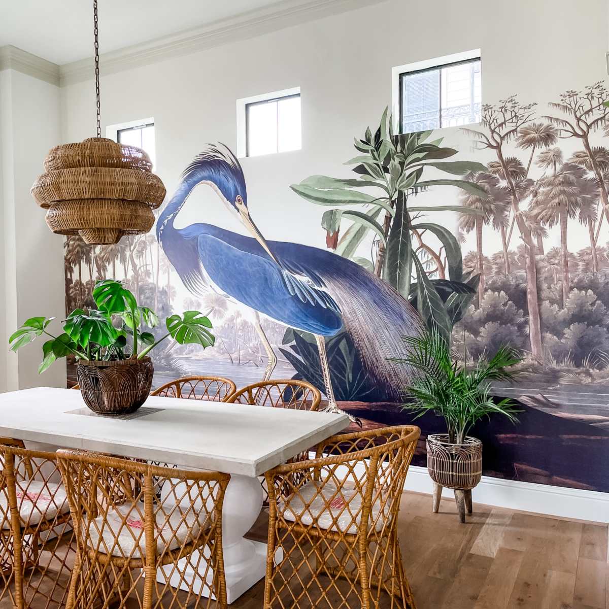 A dining room features a large mural of a blue heron and a table surrounded by wicker chairs with a potted plant in the center.