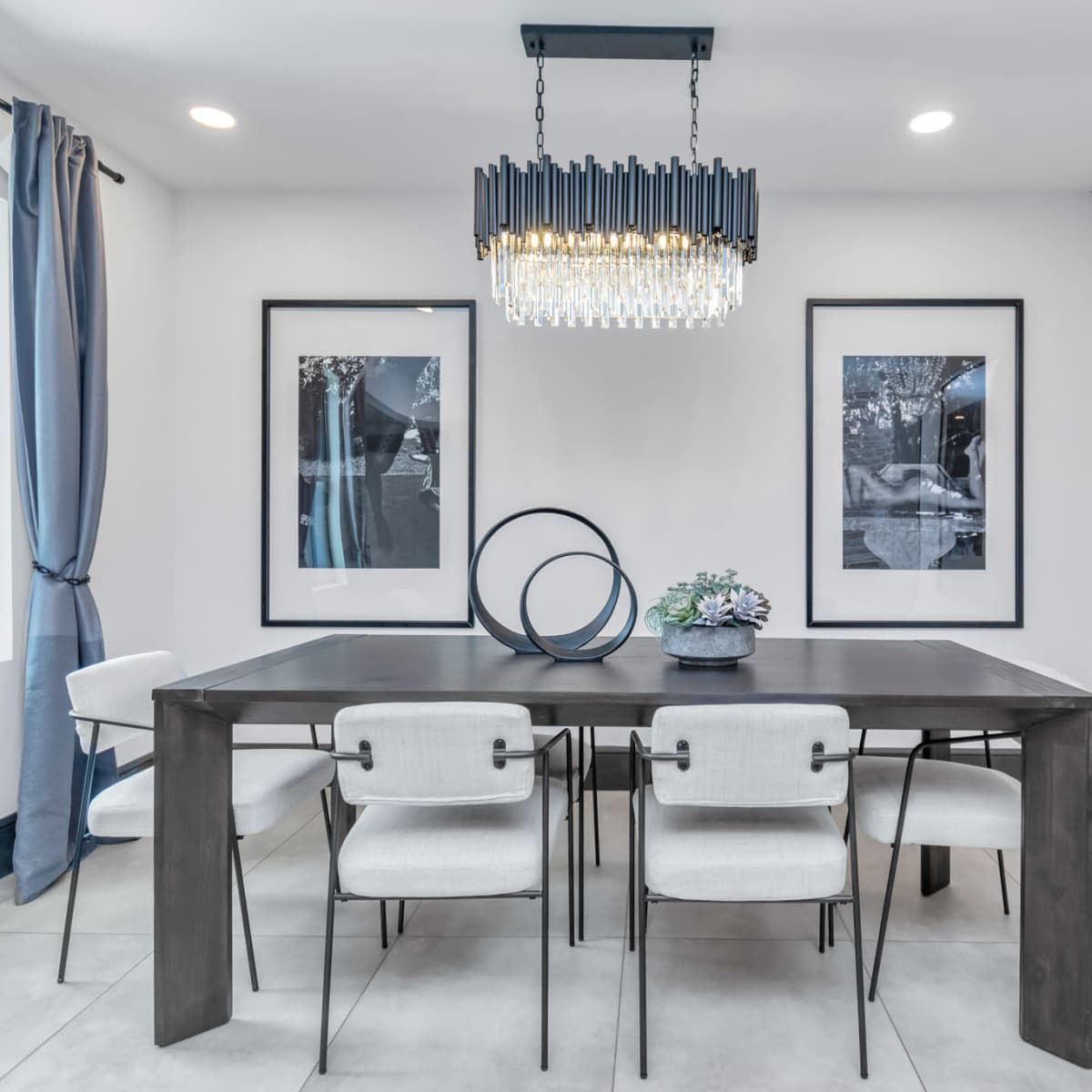 A modern dining room features a rectangular table surrounded by white upholstered chairs, with framed black-and-white photographs on the walls and a chandelier overhead.