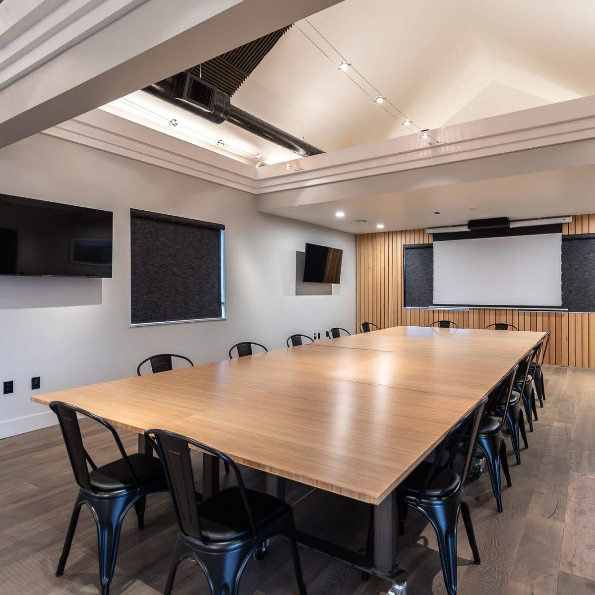A spacious conference room with a large wooden table, black chairs, and a screen at the front, complemented by wall-mounted TVs.