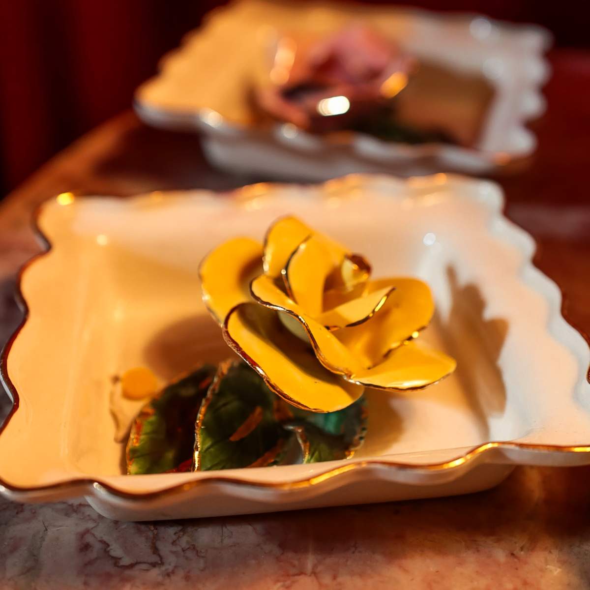 Two decorative ceramic dishes, one featuring a yellow flower and the other a pink flower, rest on a table with a red backdrop.