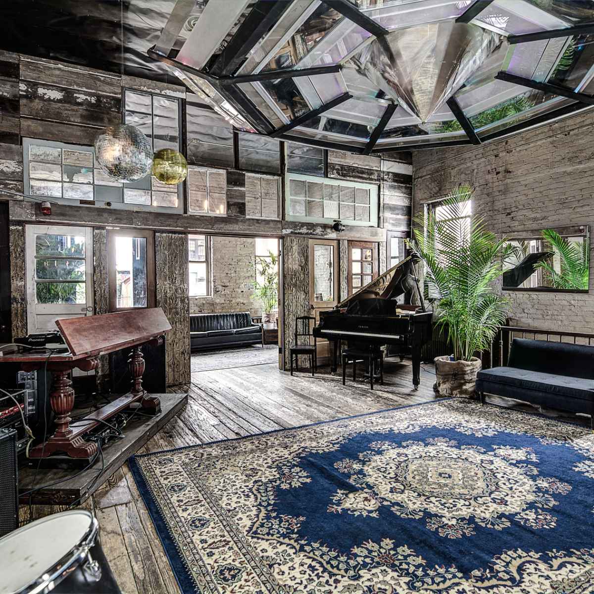 A spacious, rustic room featuring a grand piano, a large area rug, various plants, and multiple windows letting in natural light.