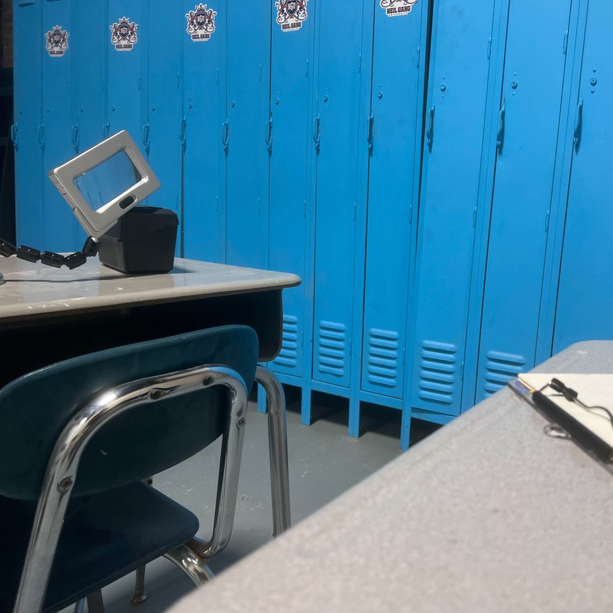 A classroom corner with blue lockers, a desk with a notebook, and a small mirror attached to a container.