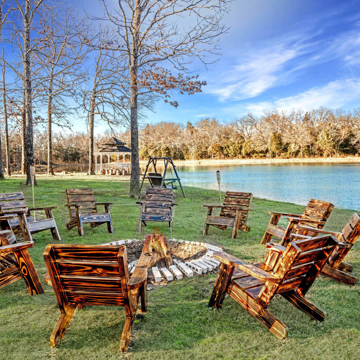 A circle of wooden chairs surrounds a fire pit near a calm lake, with trees and clear blue skies in the background.