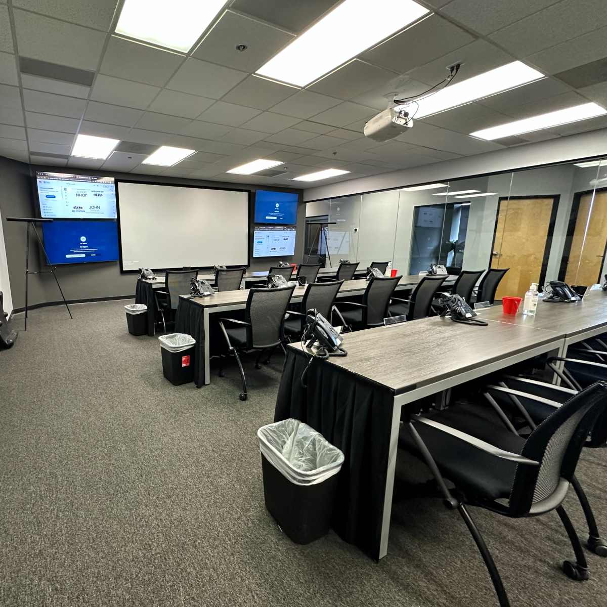 A conference room with multiple black chairs arranged around a long table, featuring two large screens and several trash bins.