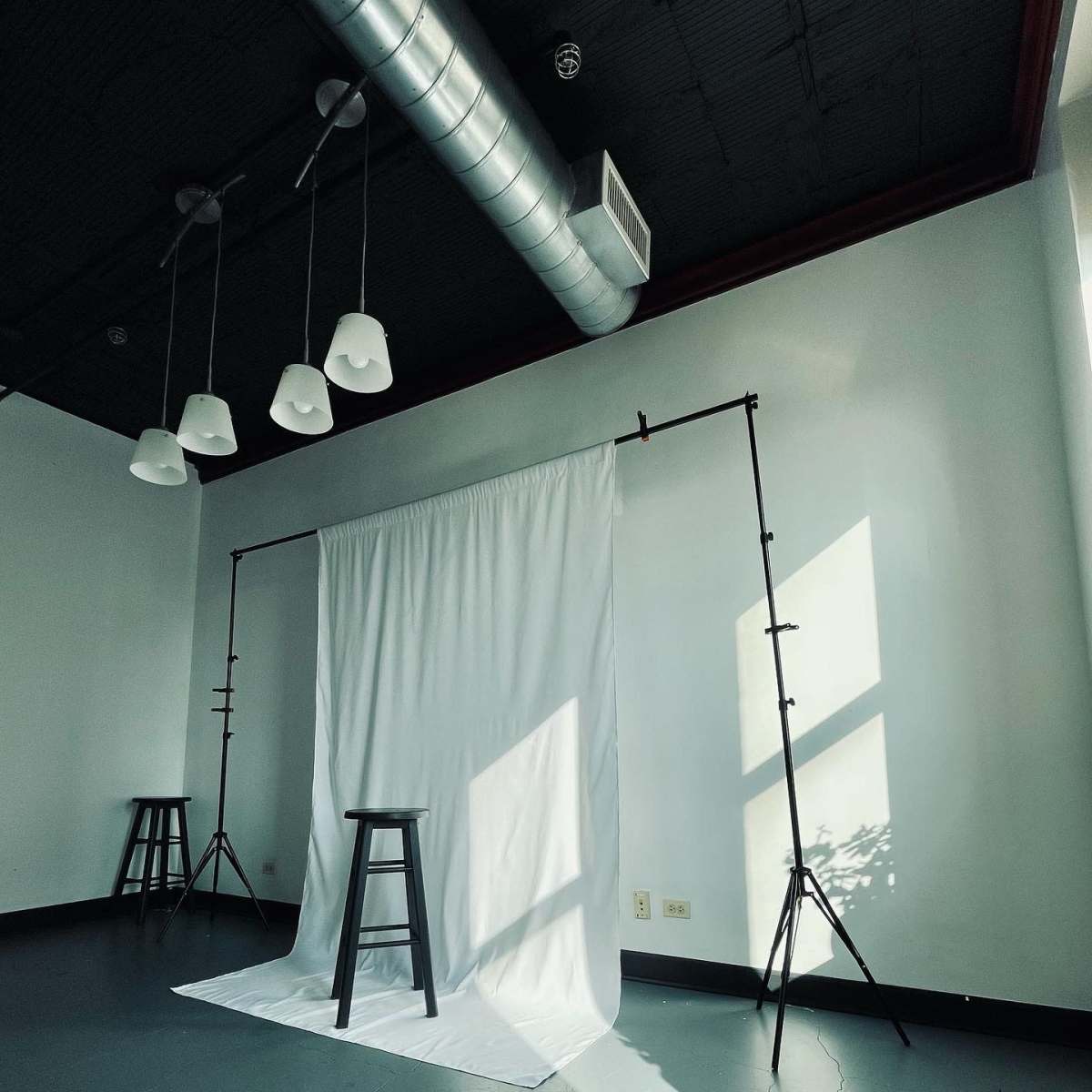 A minimalist studio space featuring a white backdrop supported by stands, with a black stool positioned in front, illuminated by natural light.