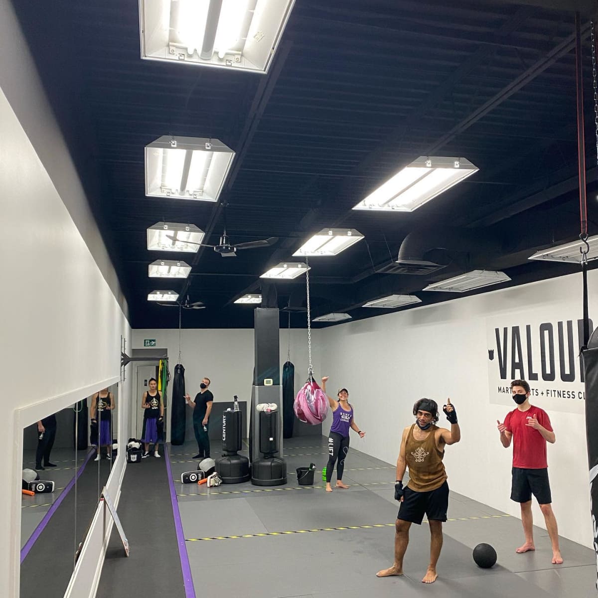 A group of people are engaged in various fitness activities inside a gym, featuring workout equipment and a boxing bag in a mirrored space.
