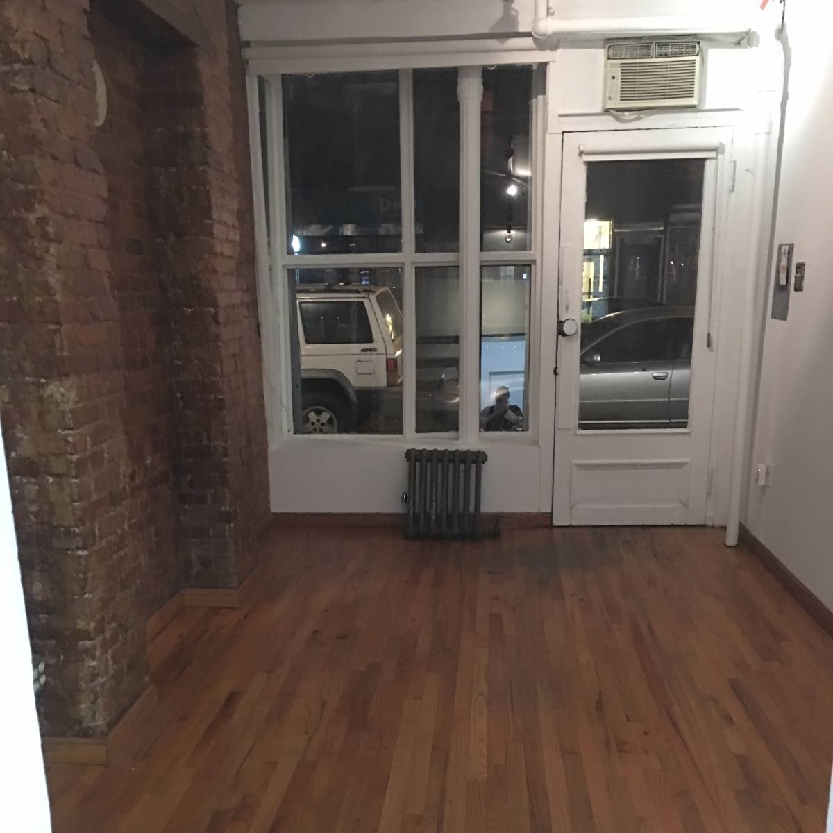 A spacious room with hardwood floors and large windows facing the street.