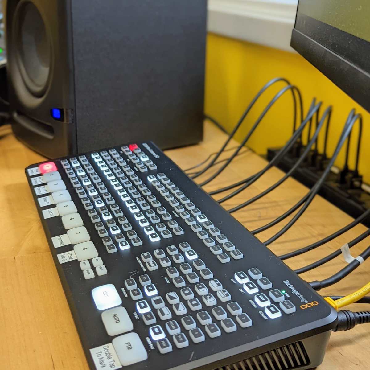 A video switcher with various buttons and a keyboard is positioned next to a speaker and several cables on a wooden surface.