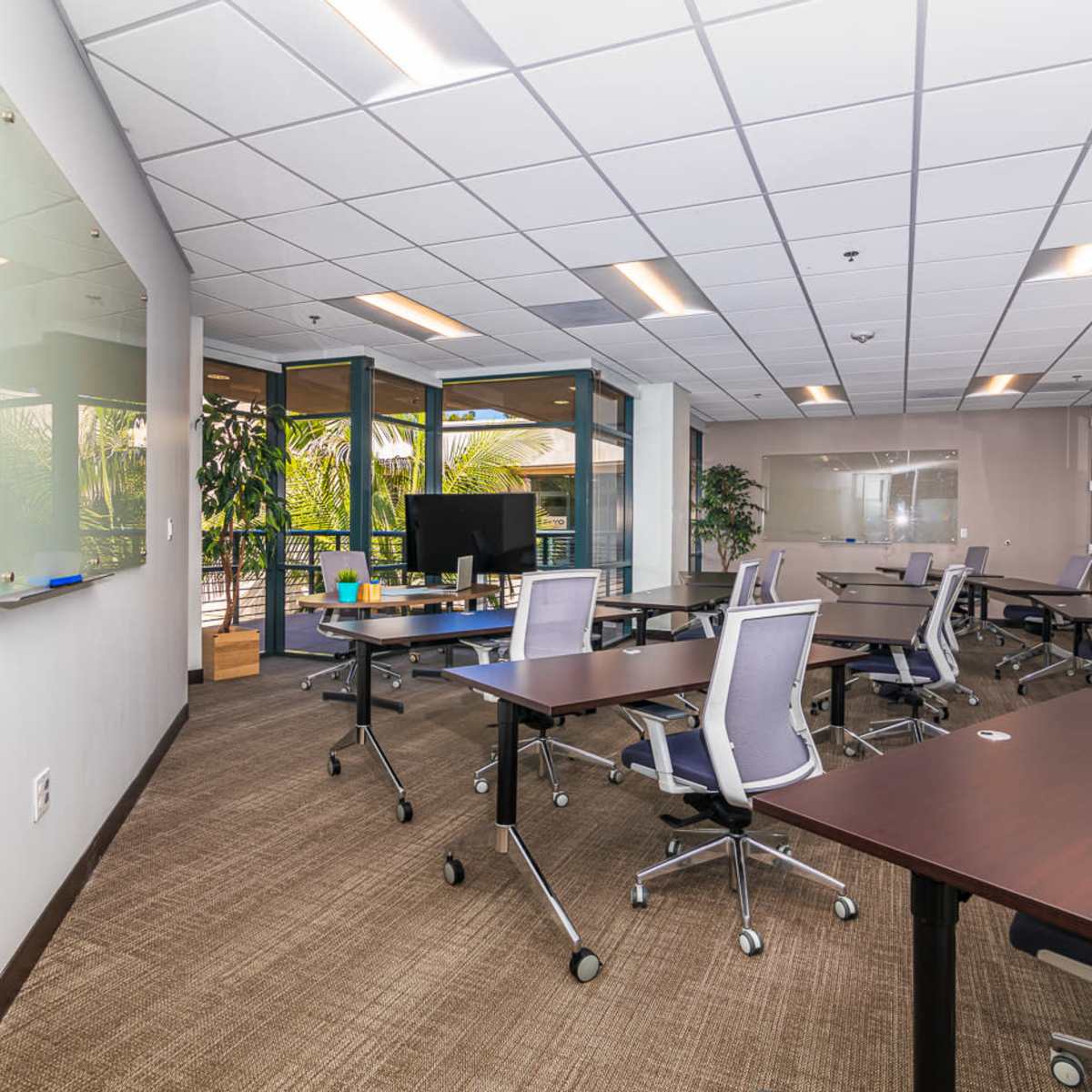 A spacious office conference room with several tables and chairs arranged in a functional layout, large windows allowing natural light, and whiteboards on the walls.