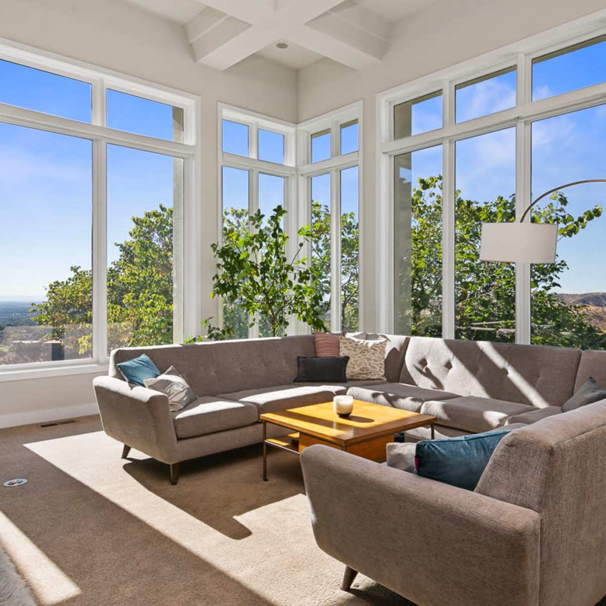 A modern living room features a large sectional sofa, a coffee table, and multiple windows offering a view of the outdoors on a sunny day.