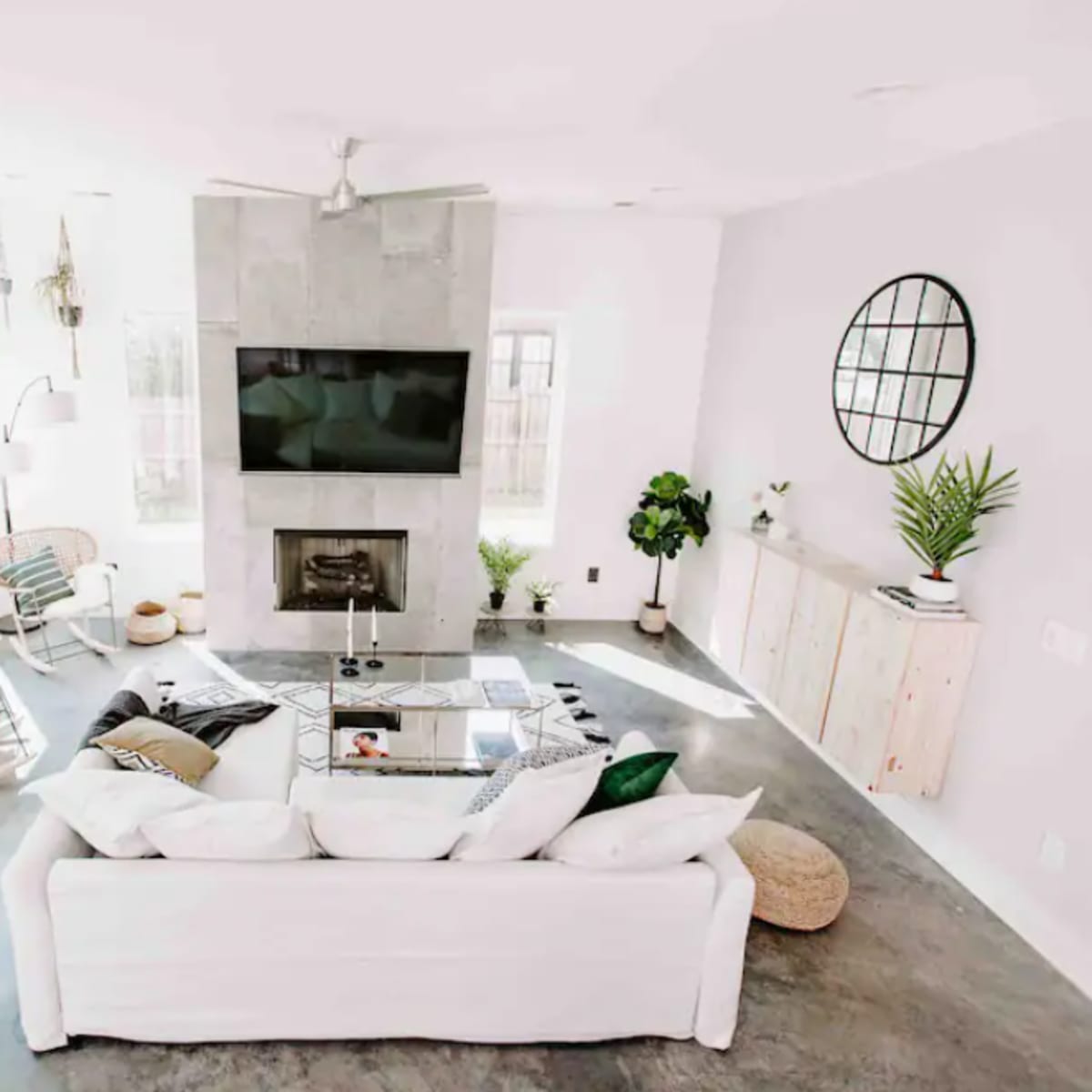 A modern living room featuring a white sectional sofa, a flat-screen TV mounted above a stone fireplace, and large windows that allow natural light to fill the space.