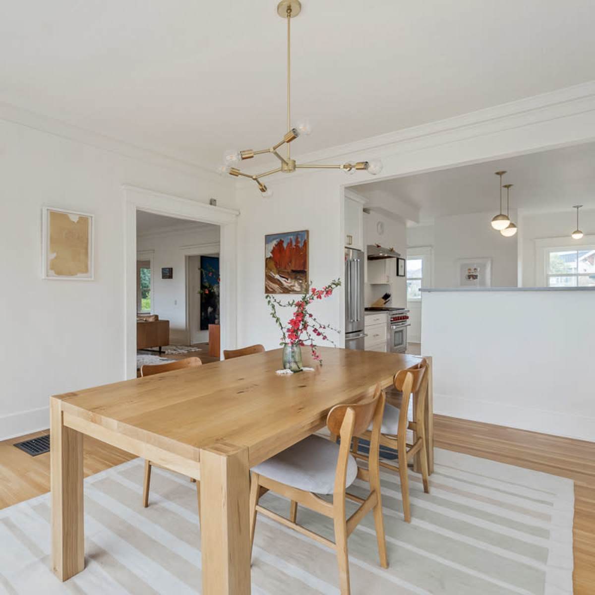 A modern dining room with a wooden table surrounded by chairs, a light fixture overhead, and an open layout leading to adjacent rooms.