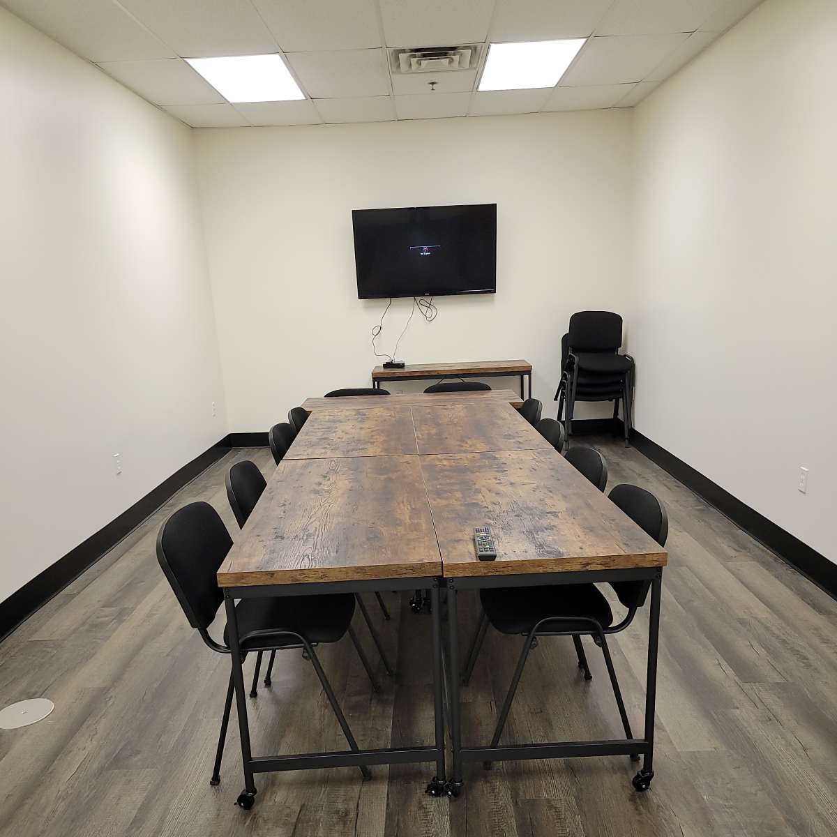 A long wooden table with chairs is set up in a minimally furnished meeting room featuring a television mounted on the wall.