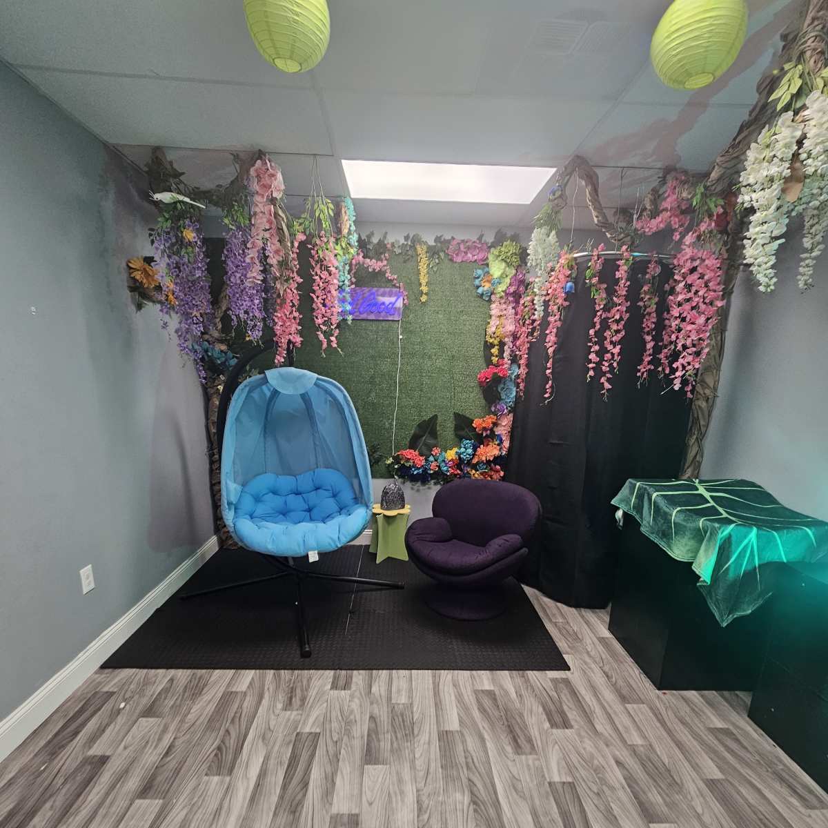 A room decorated with artificial flowers, green foliage, and colorful hanging lanterns, featuring a blue hanging chair and a purple chair on a wood-like floor.