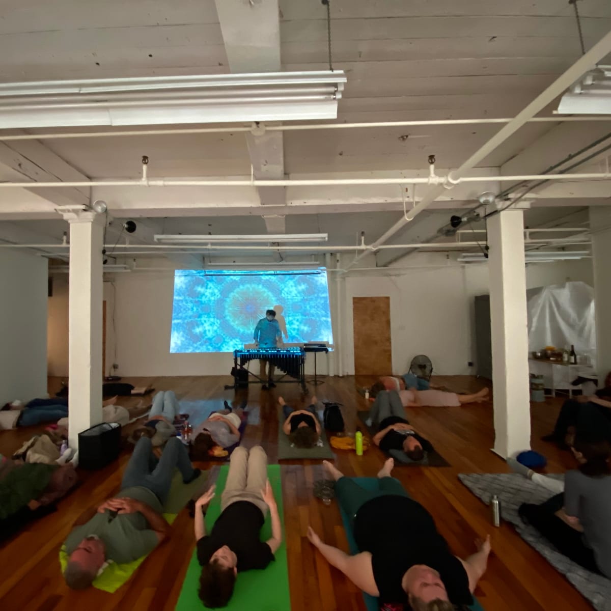 A group of people lies on yoga mats in a spacious room while a person plays a musical instrument in front of a large, colorful projection.