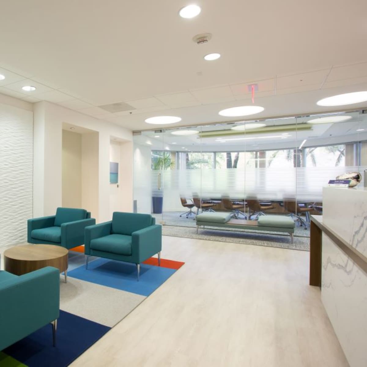 A modern office lobby with teal chairs arranged around a small wooden table, adjacent to a conference room with large windows.