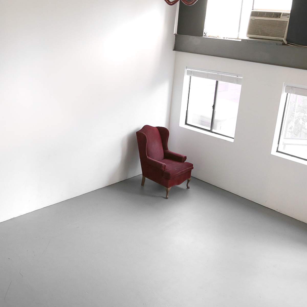 An empty room features a single red armchair positioned against a white wall, with two windows allowing natural light.