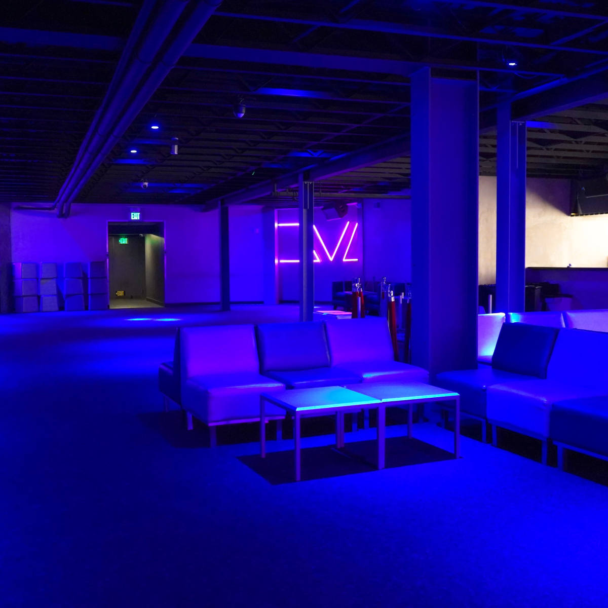 A dimly lit lounge area with blue lighting, featuring several white couches and a bar in the background.