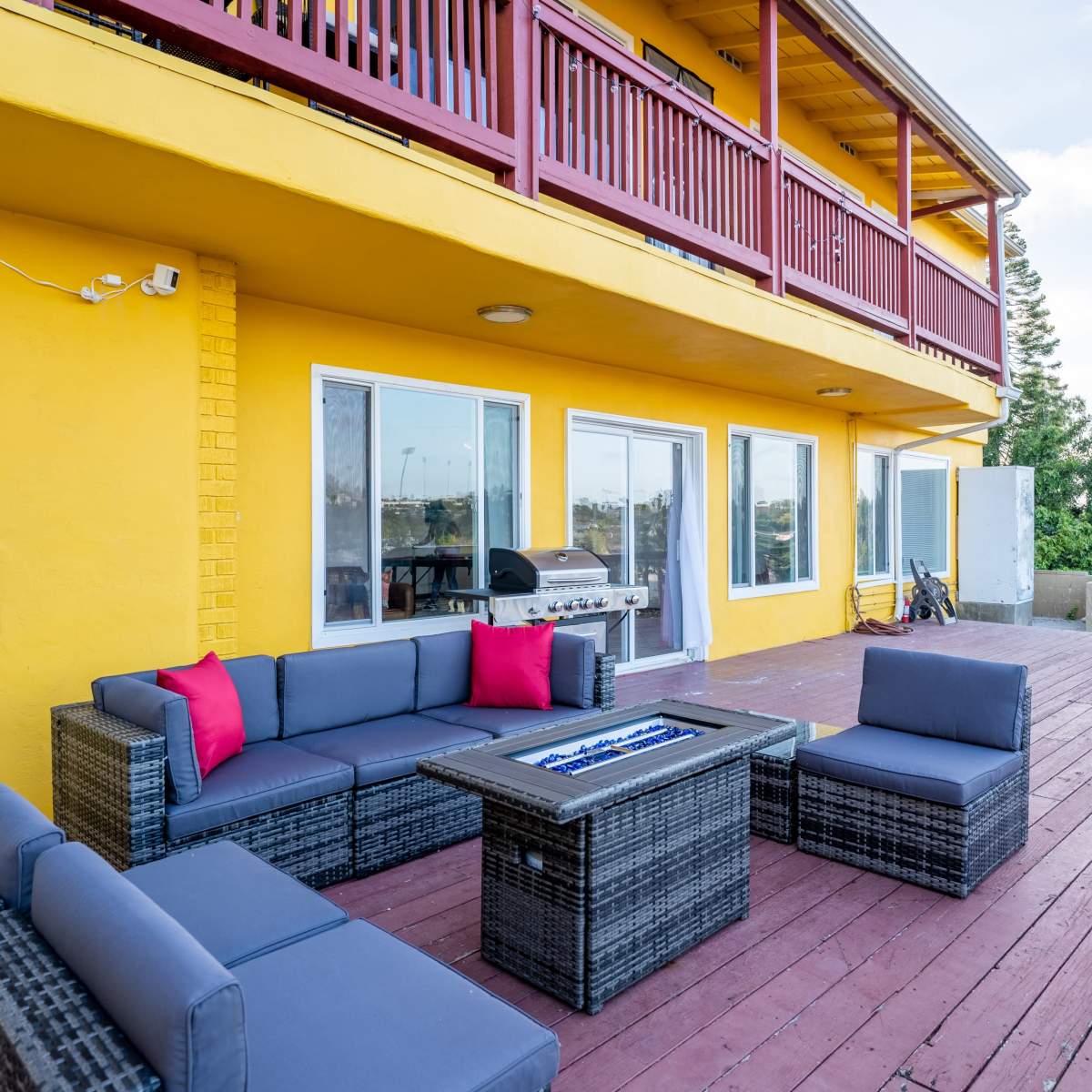 A deck with a seating area consisting of a dark wicker sofa set and a fire pit, adjacent to a yellow building with a second floor balcony.