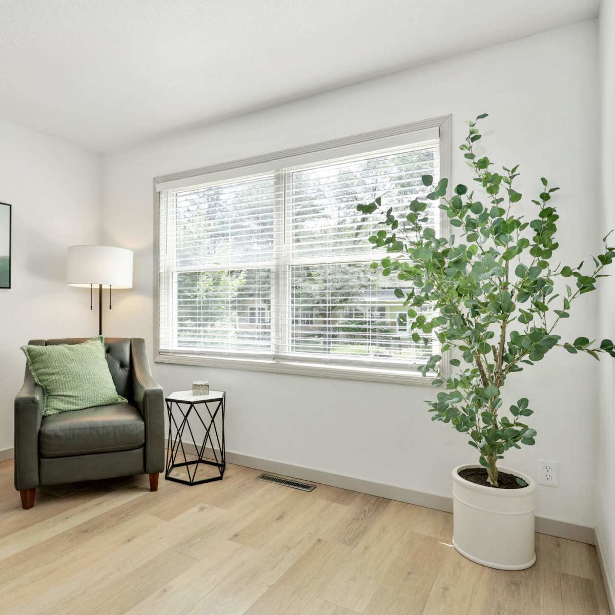 A cozy corner of a room features a green armchair beside a small black side table, a white planter with a plant, and a large window with blinds.