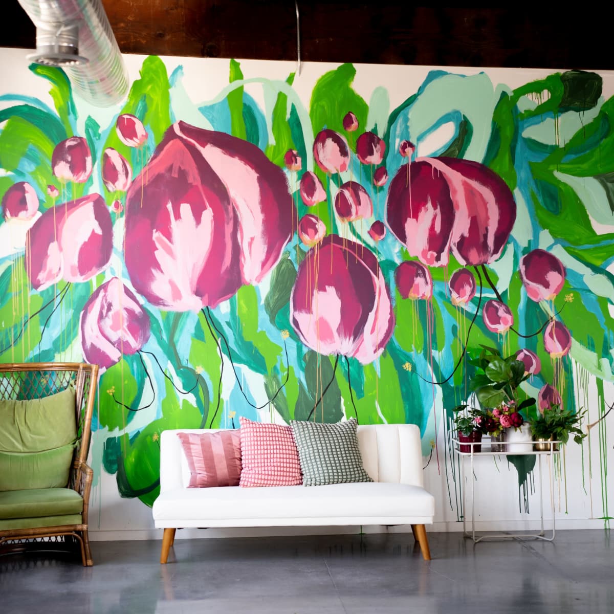 A bright interior with a large floral mural on the wall, a white sofa, a colorful pillow arrangement, and a green chair.