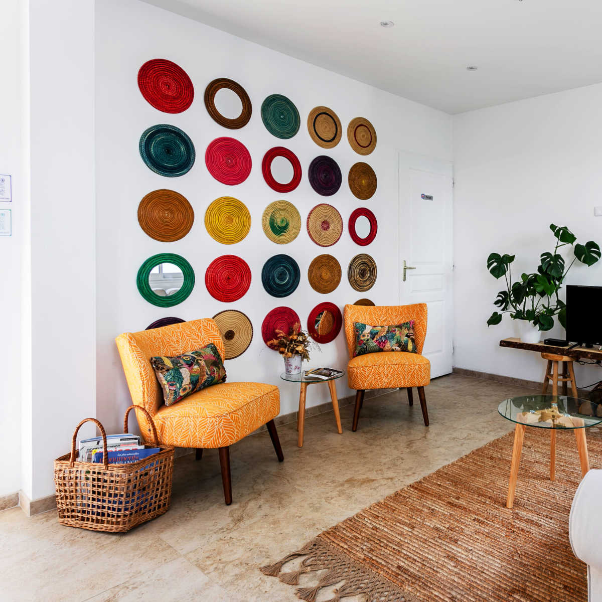 A bright living room with two orange chairs, a white sofa, a coffee table, a television, and a wall adorned with colorful decorative plates.