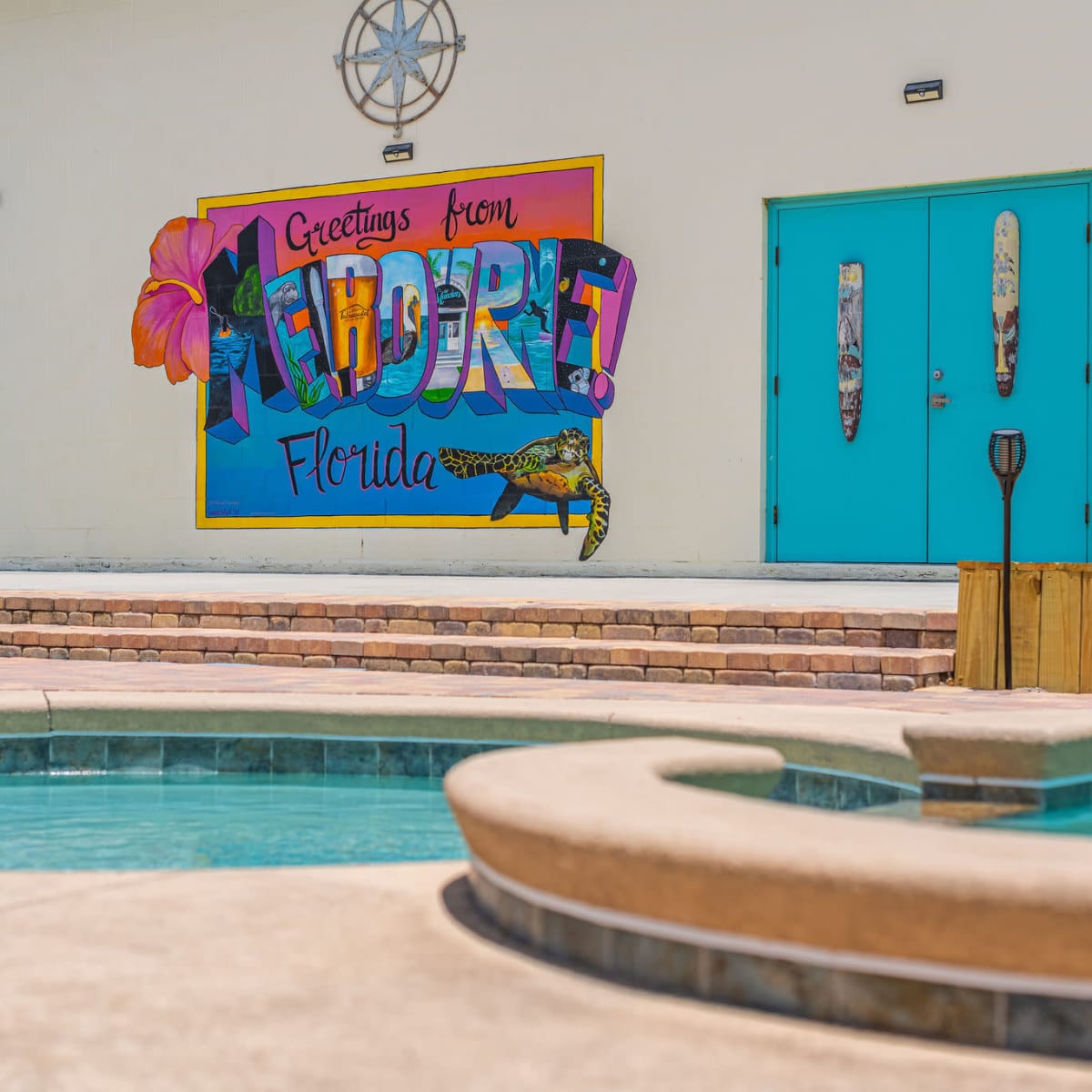A colorful mural reading "Greetings from Florida" is displayed on a wall near a swimming pool area with a hot tub, basketball hoop, and turquoise doors in the background.