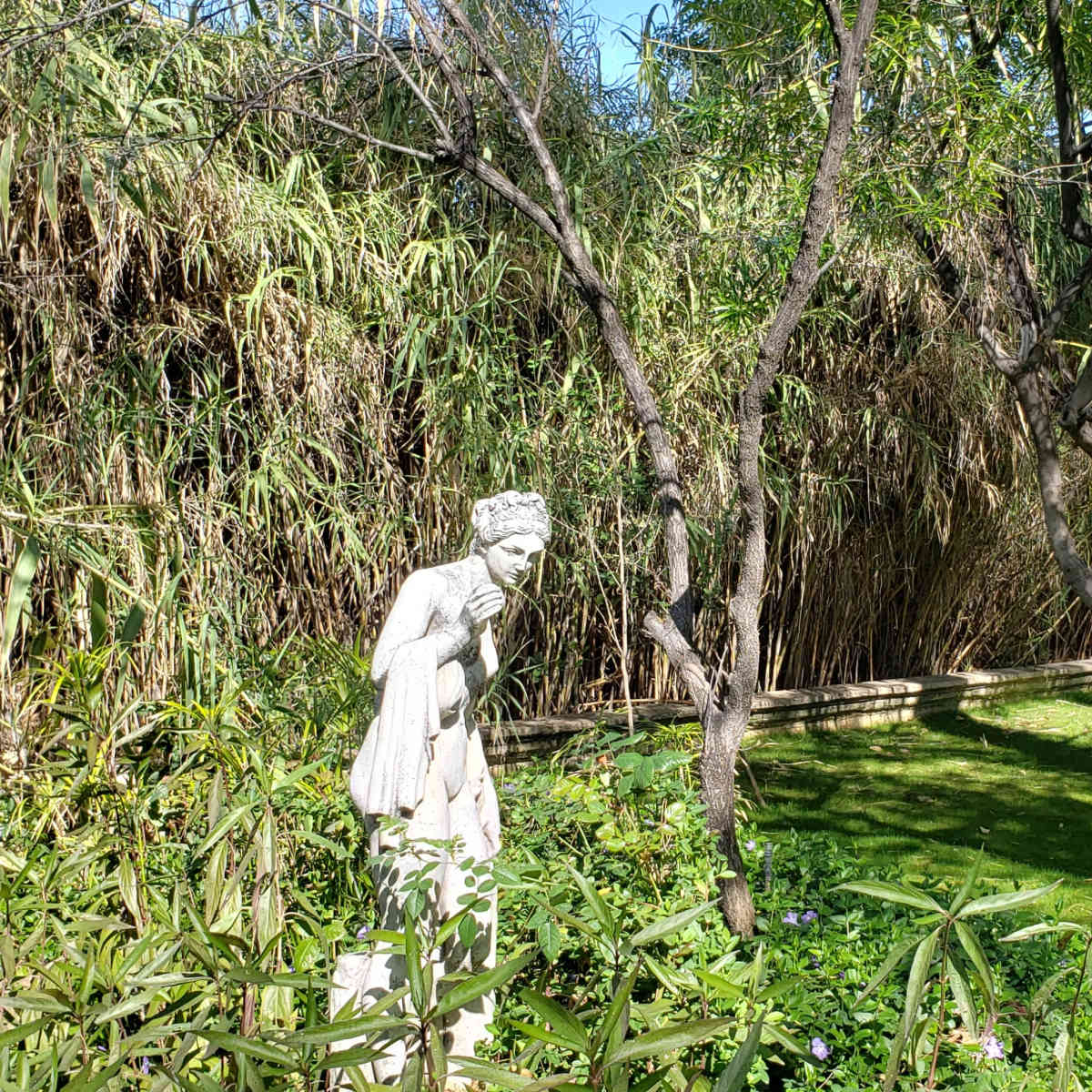 A white statue stands amidst tall grass and trees in a green garden.