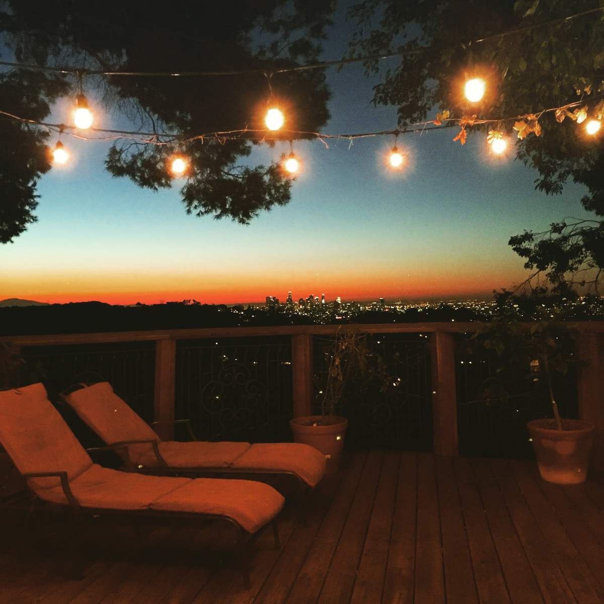 Two lounge chairs are positioned on a wooden deck with string lights overhead, overlooking a city skyline at sunset.