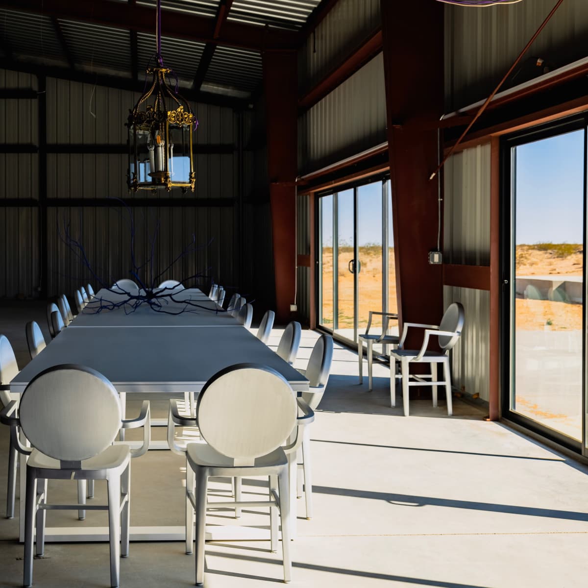 A long, rectangular dining table with chairs is set in a spacious, industrial-style room featuring large windows and a pendant light fixture.
