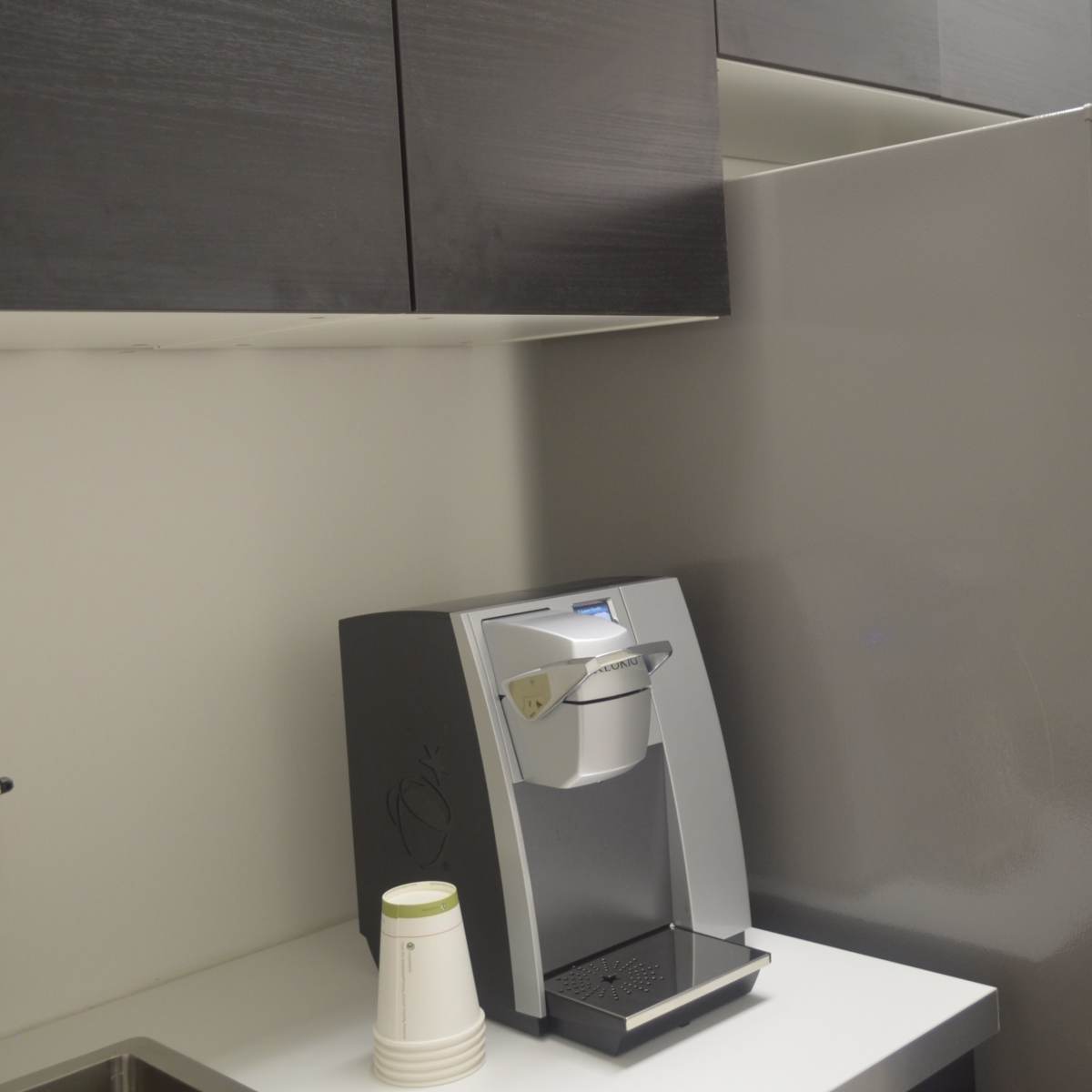 A modern kitchenette with a coffee maker on a white countertop next to a stainless steel refrigerator and black cabinet above.