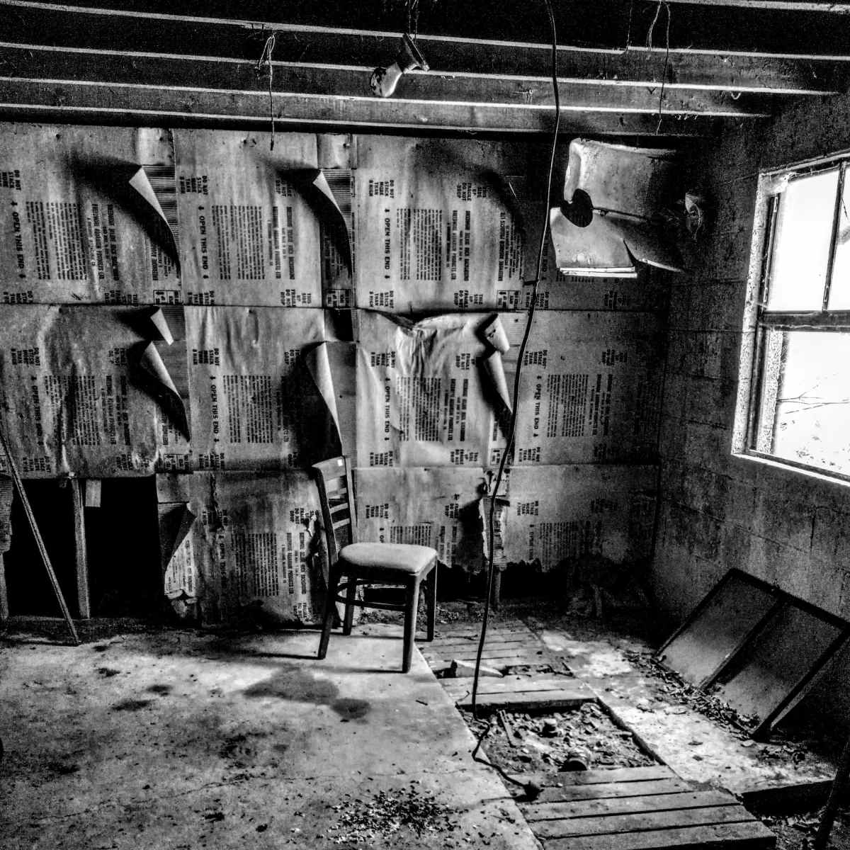 A single wooden chair stands in a dimly lit, abandoned room with bare concrete walls and scattered debris on the floor.