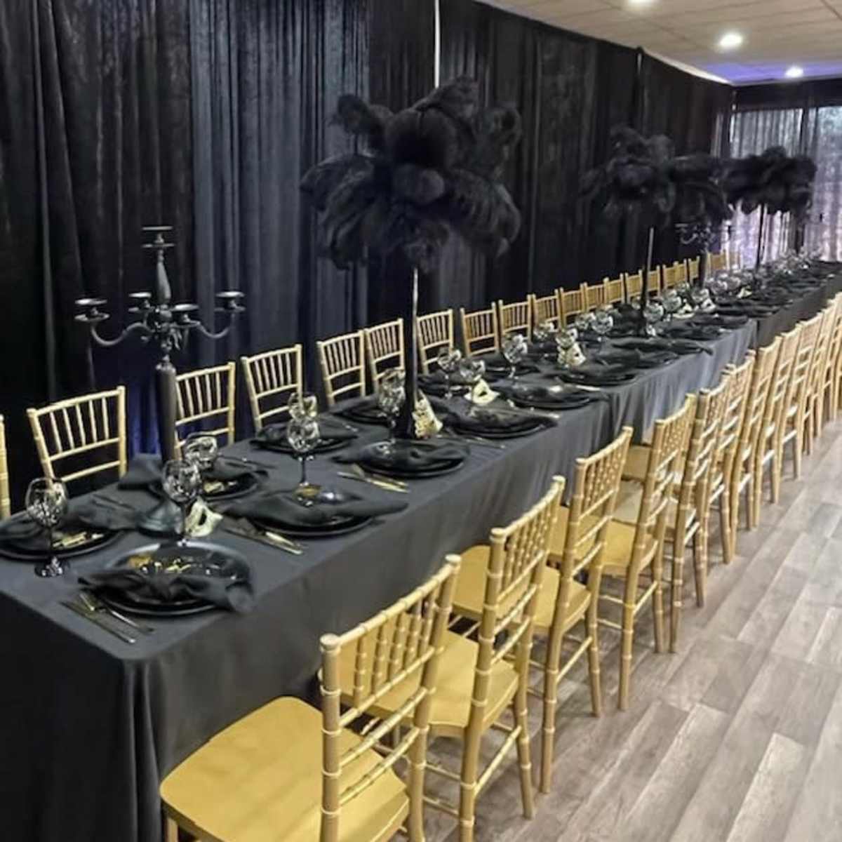 A long banquet table is set with black tablecloths, elegant gold chairs, and tall black feather centerpieces.