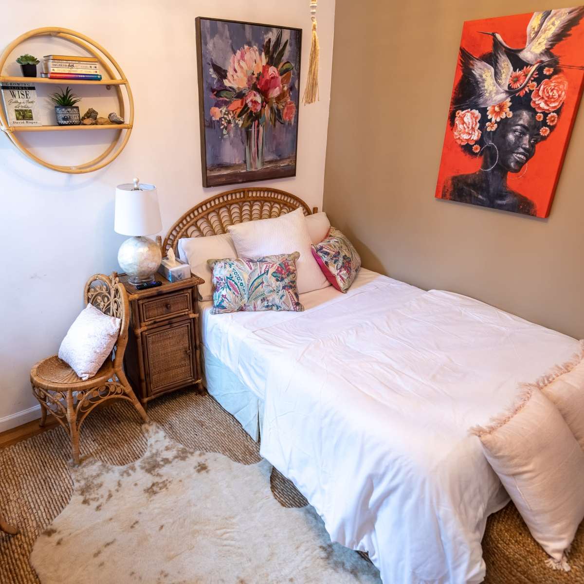 The room features a bed with white bedding, a rattan chair, a small glass-topped table, and colorful artwork on the walls.