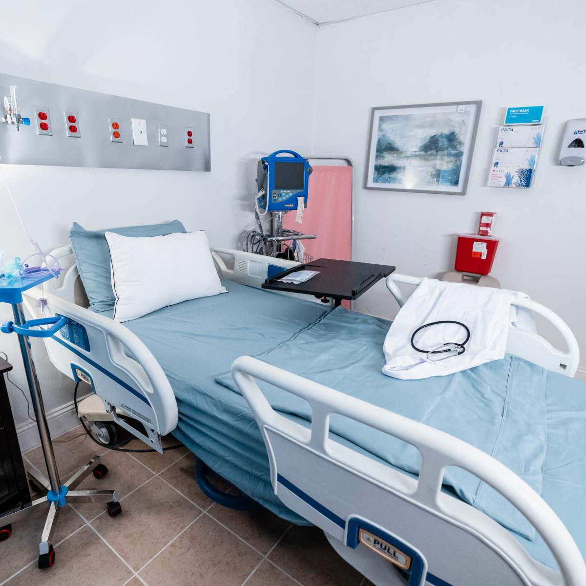 A hospital room featuring a patient bed with light blue linens, medical equipment, and a small bedside table with a lamp.