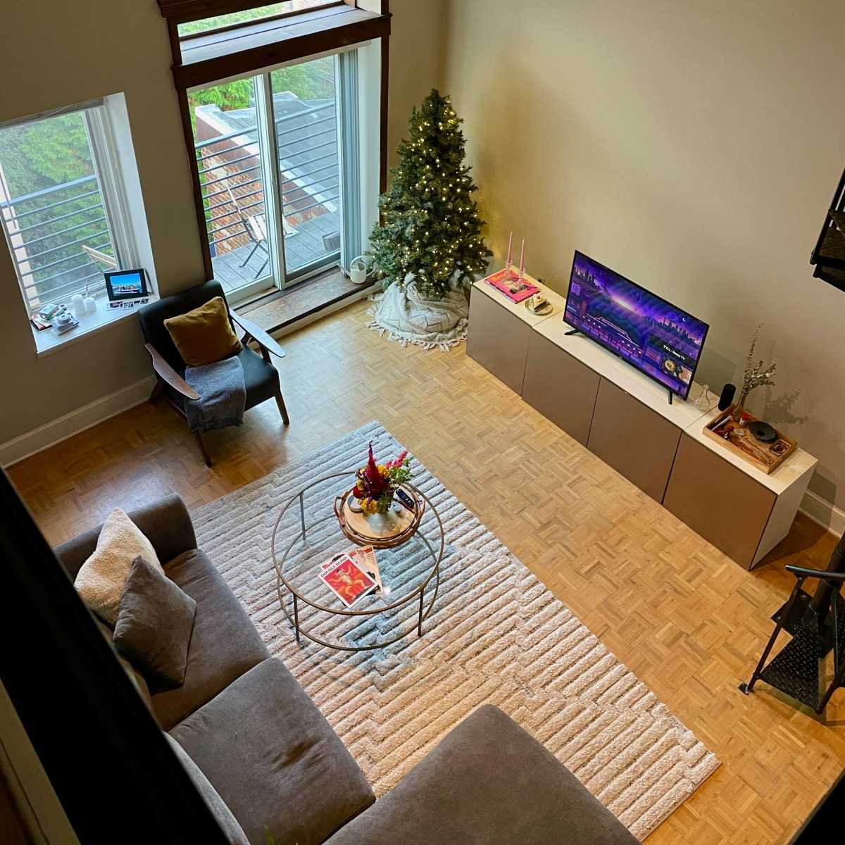 A neatly arranged living room featuring a Christmas tree, a television, a coffee table with decorations, and large windows that open to a balcony.