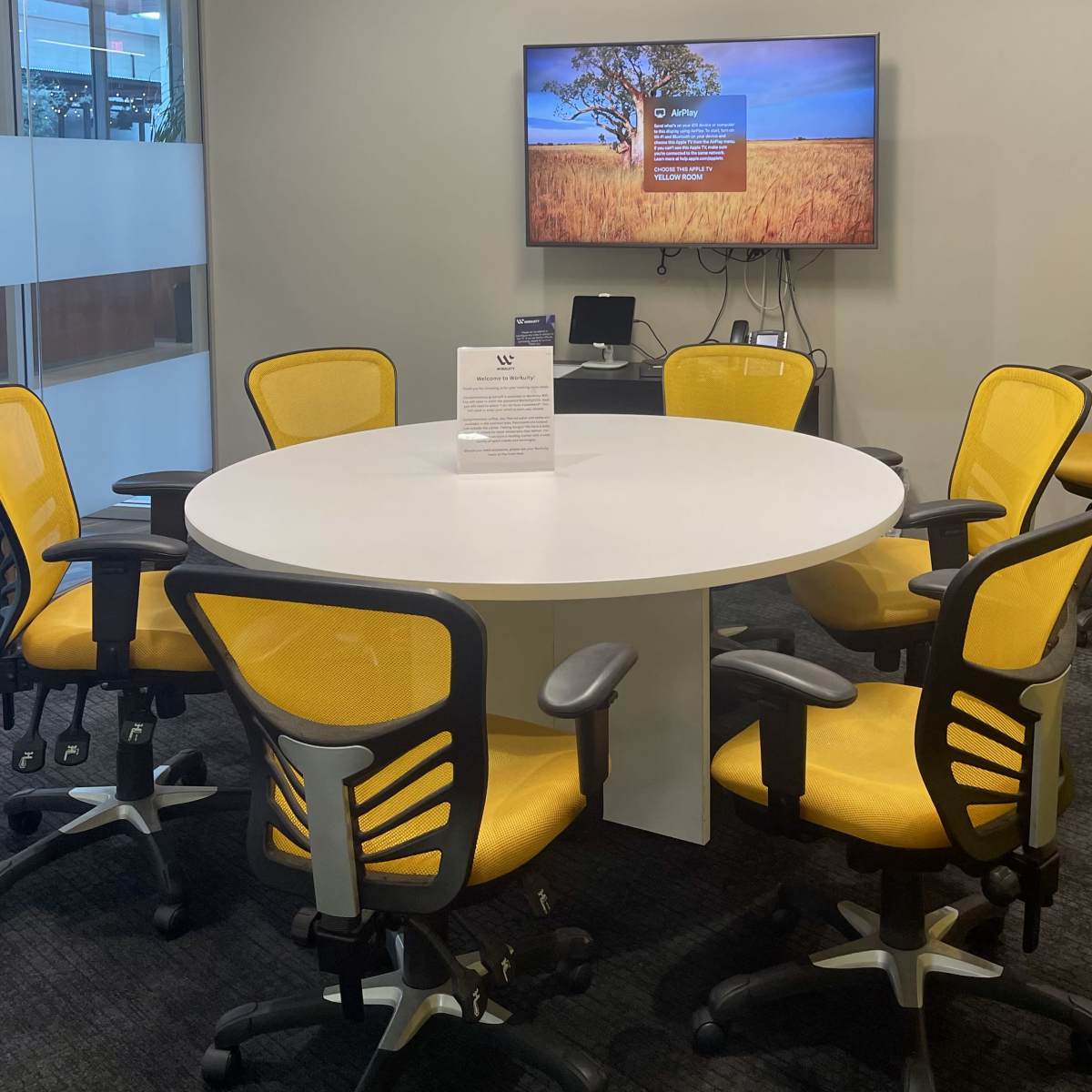 A modern meeting room features a circular white table surrounded by six yellow chairs and a wall-mounted TV displaying a landscape image.