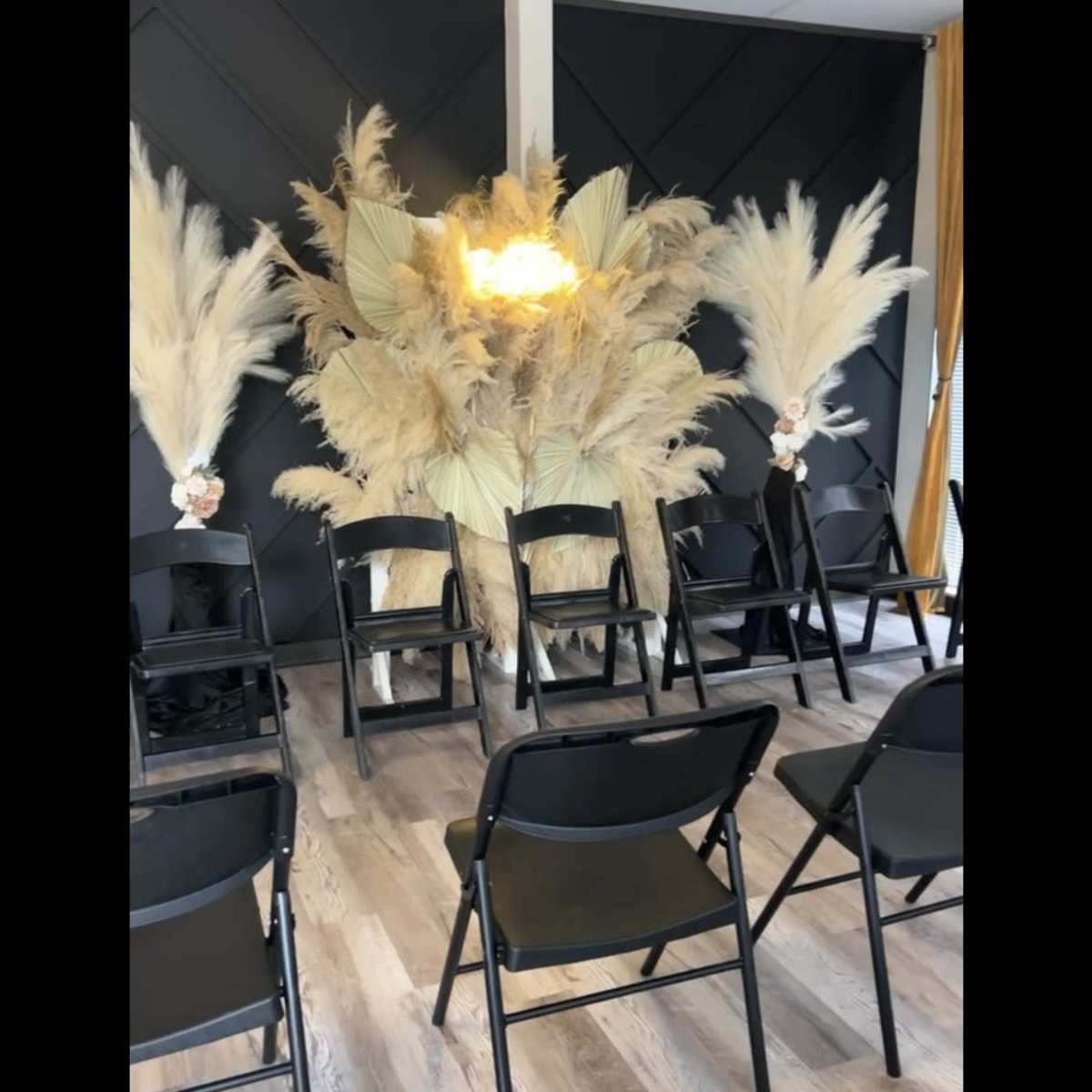 A set of black chairs is arranged in front of a large decorative backdrop made of pampas grass and a light fixture.