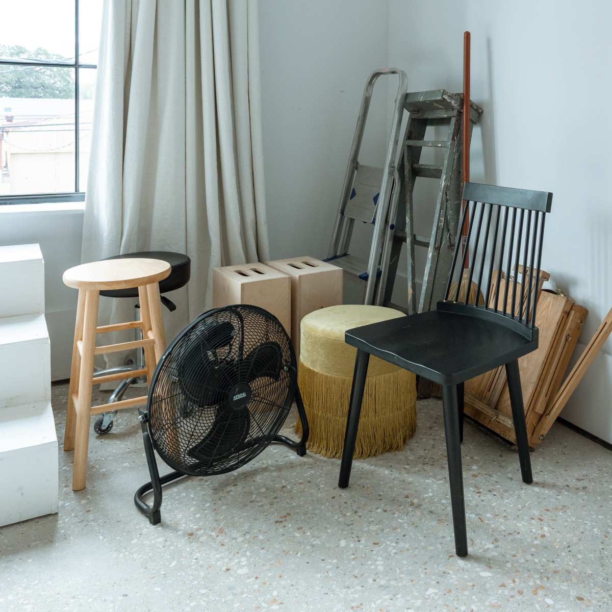 A corner of a room features two chairs, a stool, a fan, a ladder, storage boxes, and a decorative pouf.