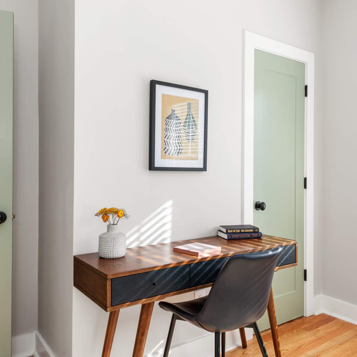 A small workspace features a wooden desk with a black chair, framed artwork on the wall, and a vase of flowers, alongside a light green door and a window with blinds.