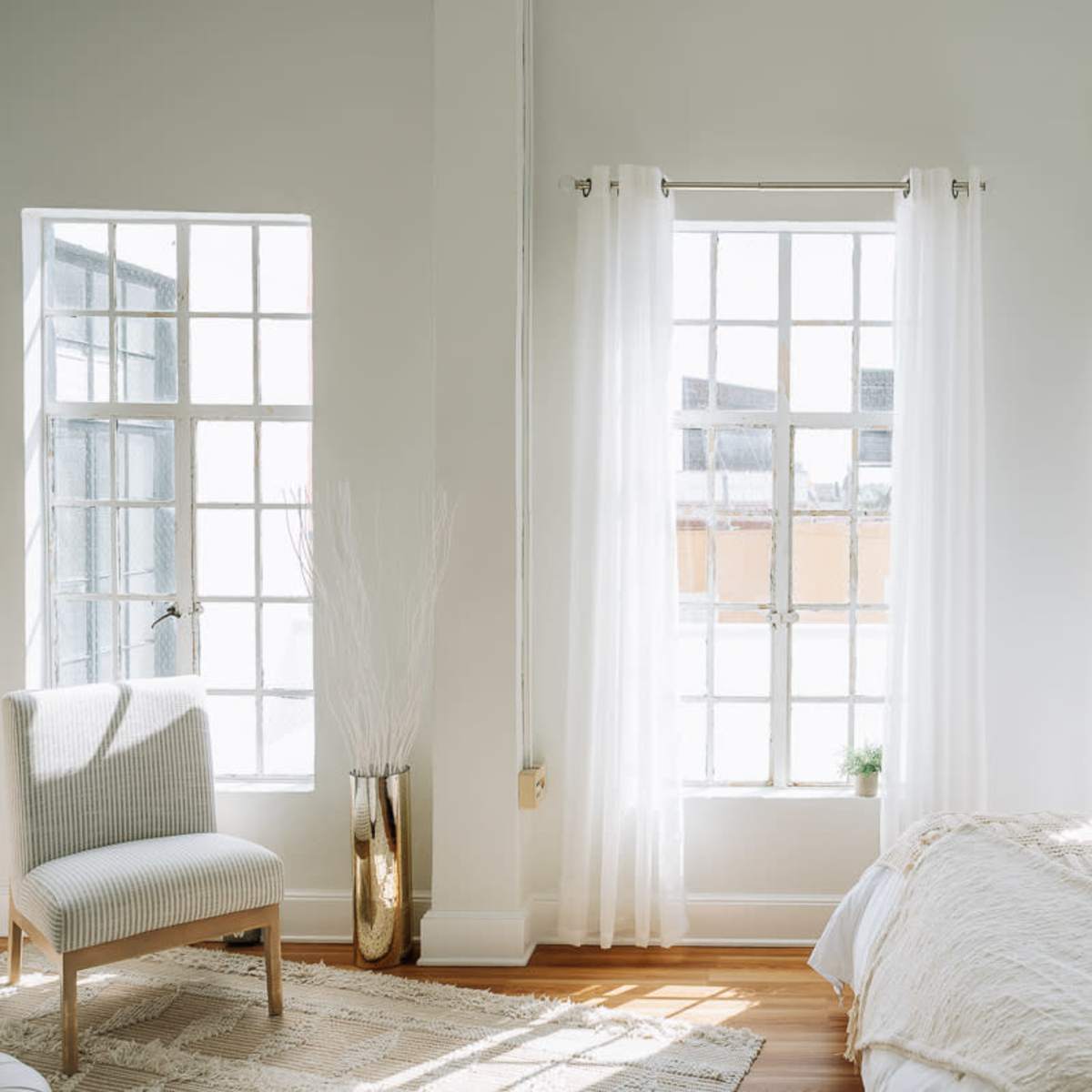 A bright bedroom features large windows adorned with sheer curtains, a bed with white linens, and a decorative chair in the corner.