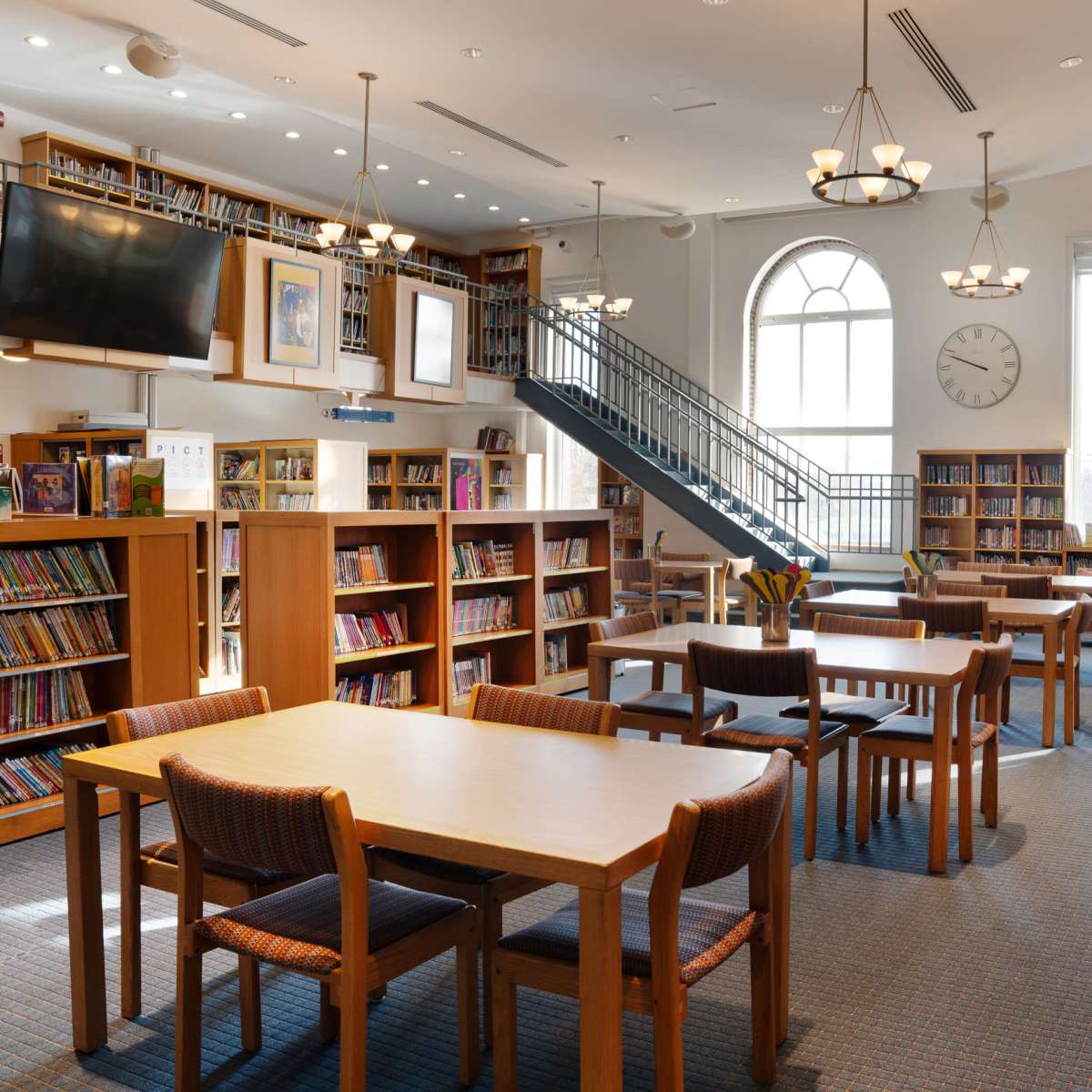 A spacious library with wooden shelves filled with books, several tables and chairs, and a staircase leading to an upper level.